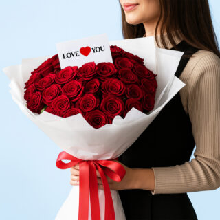A woman holding a large bouquet of vibrant red roses with a small card that says "LOVE YOU”. The flowers are wrapped in white paper and tied with a red satin ribbon.