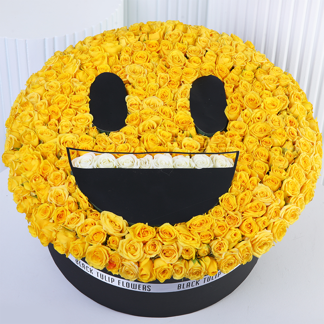 smiley-yellow-rose-box Floral arrangement shaped like a yellow grinning emoji face, made with yellow roses, black round eyes, a wide black smile, and white roses representing teeth.