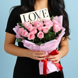 A bouquet of 16 light pink roses wrapped in pink paper with a red ribbon, with a message card with the word "LOVE" is tucked into the flowers.