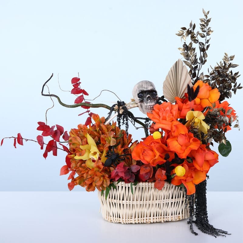 Halloween basket flower arrangement with orange roses, alstroemeria, yellow cymbidium orchids, dried leaves, and spooky Halloween decor.