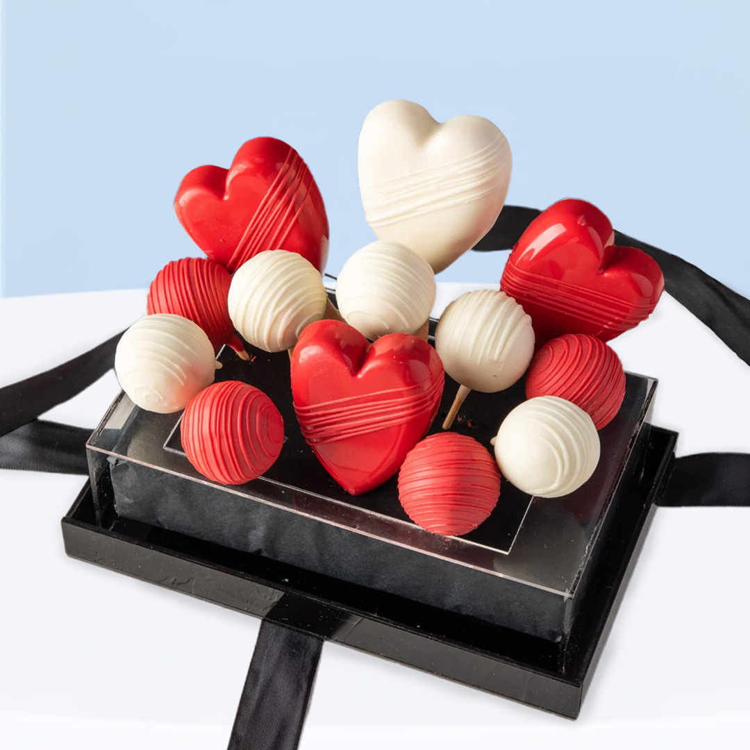 cakesicle-truffle-gift-box_01 Red and white heart-shaped cakesicles and assorted chocolate truffles in a clear gift box with black ribbon.