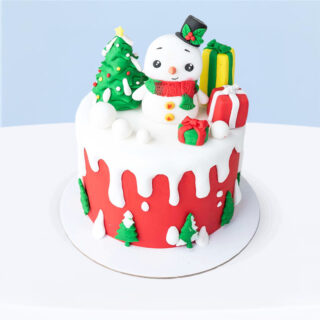 Cute red and white Christmas cake with fondant snowman, Christmas trees, and gifts inside a clear box with red ribbon.