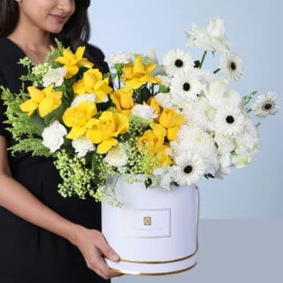 A white round box containing yellow roses, white roses, white carnations, white gerbera daisies, baby white orchids, solidaster flowers, and gypsophila arranged with decorative foliage in a bright and elegant floral arrangement.