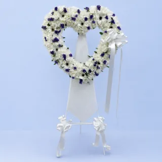 Heart of Remembrance Stand featuring white chrysanthemums, white gerberas, white tanacetum, blue statice, and fresh greenery arranged in a heart-shaped sympathy flower stand on a white steel stand.