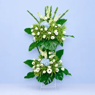 Peaceful Tribute Stand featuring white roses, white gerberas, blue hydrangeas, white matthiola, safari sunset, and lush greenery arranged on an elegant white steel sympathy flower stand.
